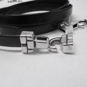May include: A black leather bracelet with two silver clasps. The clasps are square with a hook on one side.