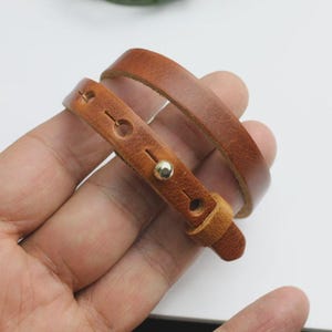 May include: A brown leather bracelet with a wrap-around design. The bracelet features a buckle closure and a gold-colored stud. The leather has a natural grain and a warm, earthy tone.