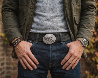 May include: A black textured leather belt featuring a silver-toned oval buckle with a raised silhouette of a cat and the text "Gran Can" embossed on the surface. The belt is worn with dark blue denim jeans, a grey heathered shirt, and an olive green jacket.