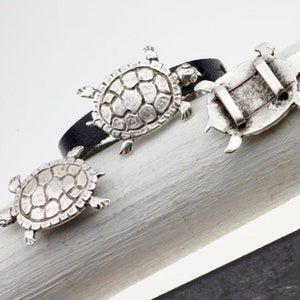 Könnte beinhalten: Ein schwarzes Lederarmband mit drei silbernen Schildkrötenanhängern. Die Schildkröten sind mit strukturierten Schalen detailliert und haben ein realistisches Design.