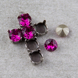 Può includere: Un ciondolo a forma di croce in metallo argentato con cinque cristalli color magenta. Sono visibili anche due cristalli sciolti, uno magenta e uno argentato. I cristalli sono rotondi e sfaccettati, riflettendo la luce.