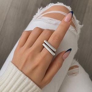 Può includere: Un anello color argento caratterizzato da due barre rettangolari orizzontali e parallele che avvolgono il dito. Il metallo ha una finitura liscia e opaca con bordi dritti e netti, creando un design geometrico che si posiziona sul dito.