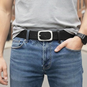 Può includere: Una cintura in pelle nera con una fibbia in metallo color argento di forma rettangolare arrotondata. La cintura è indossata su un paio di jeans blu, abbinata a una t-shirt grigio chiaro tinta unita e un orologio digitale nero al polso.