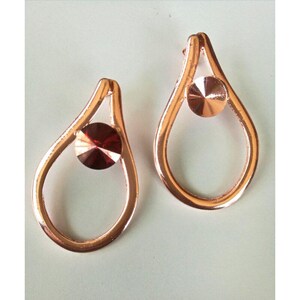 May include: A pair of rose gold teardrop earrings. Each earring features a teardrop-shaped frame with a round, faceted, dark red gemstone set in the center. The earrings have a simple, elegant design, suitable for everyday wear or special occasions.