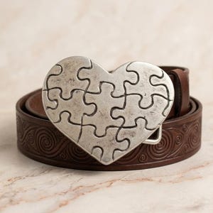 Könnte beinhalten: Ein brauner Ledergürtel mit einer silbernen herzförmigen Schnalle. Die Schnalle besteht aus Puzzleteilen. Der Gürtel hat ein dekoratives Wirbelmuster.
