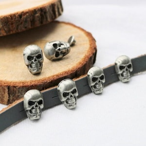 May include: Six silver skull-shaped metal studs, some attached to a gray leather strap. The studs have a detailed design and are perfect for adding a punk rock or gothic touch to clothing or accessories.