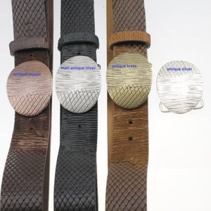 Op de afbeelding: Vier leren riemen met verschillende ronde metalen gespen in verschillende kleuren. De gespen zijn gelabeld met "antiek koper", "mat antiek zilver", "antiek messing" en "antiek zilver".