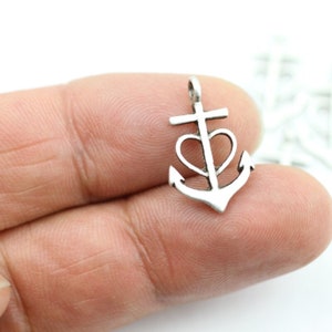 Puede incluir: Un pequeño dije de ancla de color plateado, con un corazón en el centro y una cruz en la parte superior. El ancla tiene un lazo para sujetarlo a una cadena o pulsera. El dije se sostiene entre dos dedos.