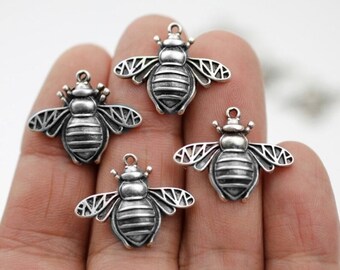 May include: Four silver-colored bee charms. Each charm features detailed wings, a segmented body, and a small loop at the top for attaching to jewelry or other crafts. The bees are arranged against a neutral background.
