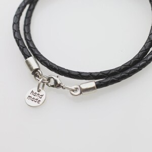 Pode incluir: Um colar de cord&atilde;o de couro tran&ccedil;ado preto com ponteiras de metal prateado, um fecho lagosta e um pequeno pingente redondo de metal prateado gravado com o texto hand made. O cord&atilde;o est&aacute; disposto em um c&iacute;rculo com as ferragens vis&iacute;veis.