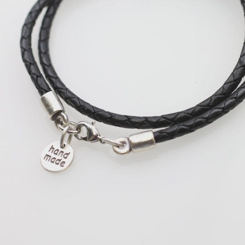 Pode incluir: Um colar de cord&atilde;o de couro tran&ccedil;ado preto com ponteiras de metal prateado, um fecho lagosta e um pequeno pingente redondo de metal prateado gravado com o texto hand made. O cord&atilde;o est&aacute; disposto em um c&iacute;rculo com as ferragens vis&iacute;veis.