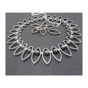 Op de afbeelding: Een zilverkleurige ketting en hanger set. De ketting heeft bladvormige schakels, elk met een donkergrijze parel. De hanger is een gestileerde bloem. De sieraden worden gepresenteerd op een donkergrijze achtergrond. De ketting is ongeveer 40 cm lang.