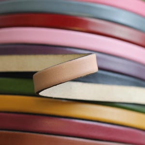 May include: A close-up of a stack of leather strips in various colors, including pink, red, green, yellow, and brown. The top strip is a light brown leather strip with a dark brown edge.