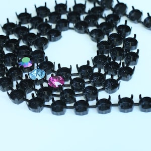 May include: A close-up of a black chain of metal settings for rhinestones. The chain is coiled and there are three rhinestones in the center of the image: one iridescent green, one light blue, and one pink.