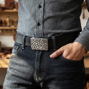 May include: A black leather belt featuring a rectangular metal buckle with a textured, raised pattern resembling scales. The belt is worn through the belt loops of dark blue denim jeans, paired with a grey button-down shirt with a visible collar.