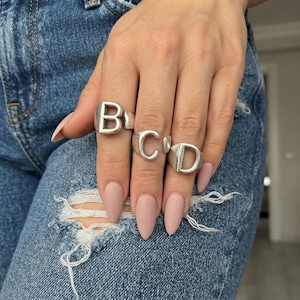 May include: Three silver-toned rings worn on fingers, each featuring a large, bold, uppercase letter. The rings display the letters B, C, and D in a smooth, metallic finish. The rings are worn on the index, middle, and ring fingers respectively.