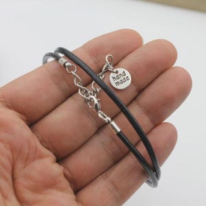 Pode incluir: Uma pulseira de cord&atilde;o de couro preto com ferragens de metal prateado, incluindo um fecho lagosta, uma corrente extensora e um pequeno pingente redondo gravado com o texto hand made em um design simples, limpo e funcional.