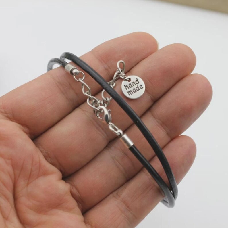 Pode incluir: Uma pulseira de cord&atilde;o de couro preto com ferragens de metal prateado, incluindo um fecho lagosta, uma corrente extensora e um pequeno pingente redondo gravado com o texto hand made em um design simples, limpo e funcional.