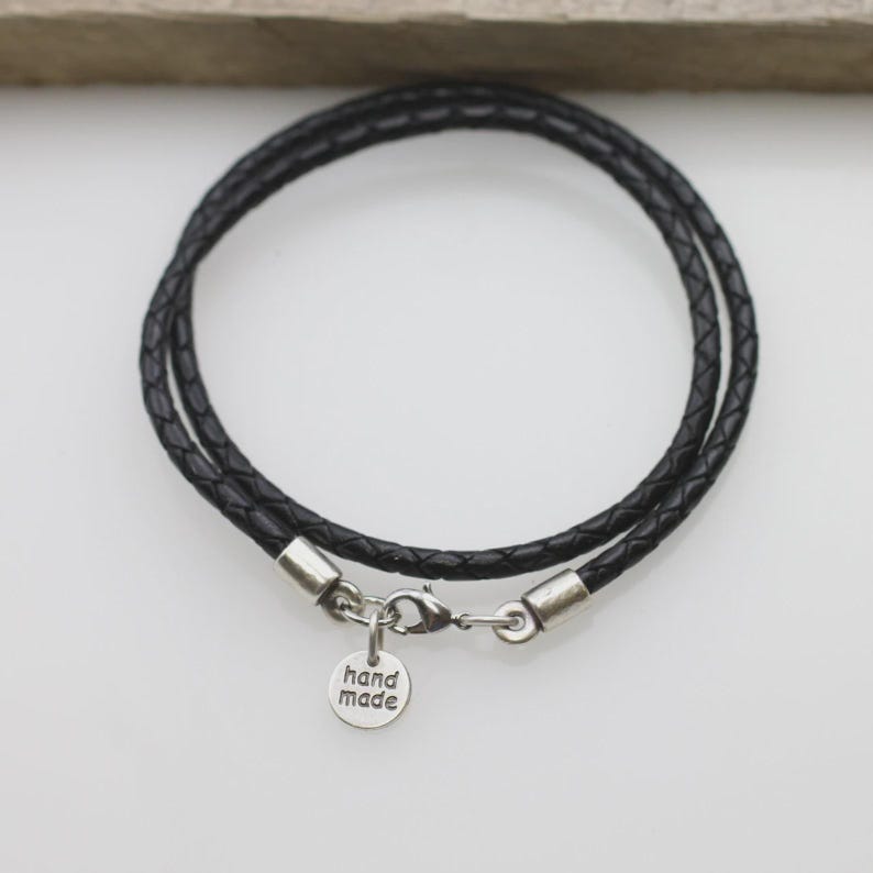 Pode incluir: Uma pulseira de cord&atilde;o de couro tran&ccedil;ado preto com um fecho lagosta prateado e um pequeno pingente redondo de metal prateado gravado com o texto hand made. A pulseira est&aacute; disposta em formato circular com extremidades sobrepostas.