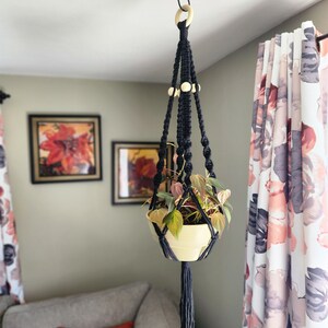 May include: A black macrame plant hanger with wooden beads hangs from the ceiling, holding a potted plant with green leaves. The hanger has a long tassel at the bottom.