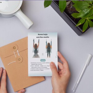 May include: A card with instructions for aerial yoga, held by a person. The card features illustrations and text in Spanish, with a brown envelope and a white pen nearby. A small plant in a black pot is also visible.
