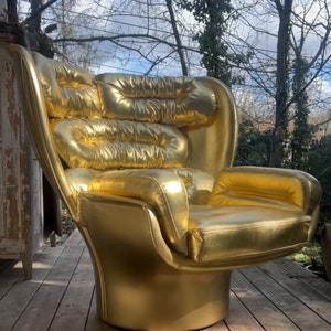 Puede incluir: Un sillón de color dorado con un diseño único, redondeado y bulboso. El sillón tiene un respaldo alto y un asiento amplio y cómodo. El sillón está hecho de un material liso y brillante que parece ser cuero.