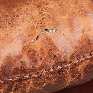 May include: Close-up of a brown leather ottoman with a stitched seam and a small tear in the leather.