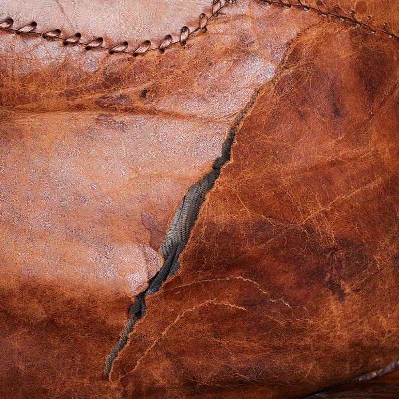 May include: Close-up of brown leather with a visible seam and a tear in the material. The leather has a distressed, vintage look.
