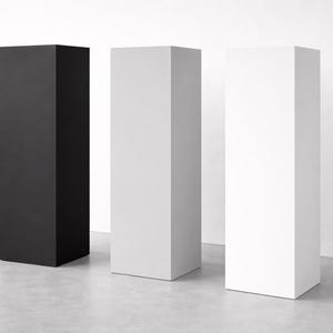 May include: Three rectangular pedestals in a studio setting. One is black, the other two are white. The pedestals are tall and narrow, with a simple, geometric design. The background is a plain white wall and a gray floor.
