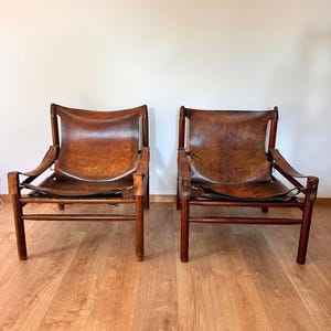 May include: Two matching lounge chairs featuring dark brown wooden frames and suspended brown leather seats. The leather is secured to the frame with visible stitching and straps, creating a low-profile seating design with a natural finish.