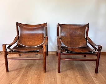 May include: Two matching lounge chairs featuring dark brown wooden frames and suspended brown leather seats. The leather is secured to the frame with visible stitching and straps, creating a low-profile seating design with a natural finish.