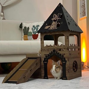 Puede incluir: Una casa de madera para conejos con rampa y techo decorativo. La casa es marrón con detalles negros y presenta un recorte de un conejo en una escoba. Un conejo esponjoso está dentro.