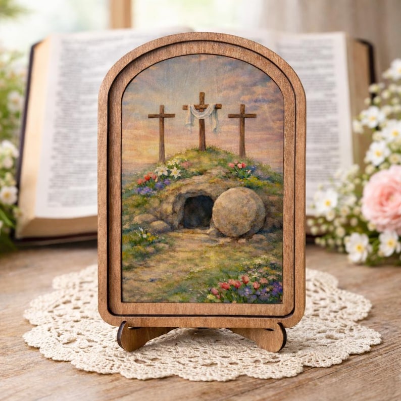 May include: An arched wooden frame containing a printed image of three crosses on a hill above a stone tomb with a rolled-away stone. The scene features a soft sky, colorful flowers, and greenery, presented as a decorative tabletop display piece.