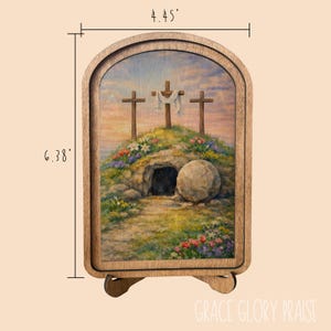 May include: An arched wooden frame containing a printed illustration of three crosses on a hill above a stone tomb with a rolled-away stone. The frame measures 4.45 inches wide by 6.38 inches high. The scene features colorful flowers and a sky.