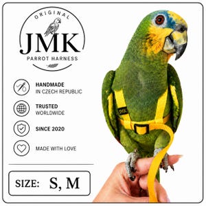 以下が含まれることがあります： 黒いバックルとJMKと書かれた小さなタグが付いた黄色いオウム用ハーネス。テキストには、ORIGINAL JMK PARROT HARNESS、HANDMADE IN CZECH REPUBLIC、TRUSTED WORLDWIDE、SINCE 2020、MADE WITH LOVE、SIZE: S, Mが含まれています。ハーネスは緑色のオウムが着用しています。