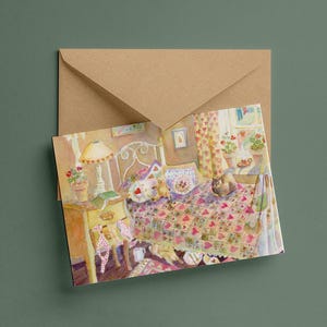 May include: A rectangular greeting card featuring a watercolour illustration of a cosy bedroom with a patterned quilt, a bedside table with a lamp, and a cat sitting on the bed, tucked into a plain light brown paper envelope for mailing.