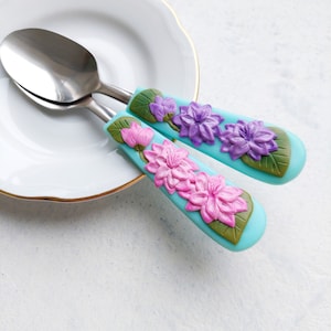 Può includere: Due cucchiai d'argento con manici blu decorati con fiori rosa e viola. I cucchiai sono su un piatto bianco.