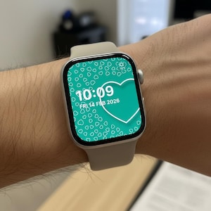 May include: A silver smartwatch with a light gray band displays a teal heart-themed watch face. The time is 10:09, and the date is Friday, February 14, 2026. The watch is on a wrist.