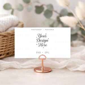 Może przedstawiać: Biała karta z napisem "Your Design Here" w eleganckiej czcionce jest trzymana przez miedziany metalowy uchwyt. Uchwyt ma okrągłą podstawę i pętlę do trzymania karty. Tło obejmuje pleciony kosz i miękką, białą tkaninę.