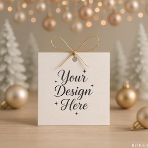 Puede incluir: Una etiqueta de regalo cuadrada, blanquecina, con el texto "Your Design Here" en escritura elegante, atada con una cinta dorada. La etiqueta se coloca sobre un fondo de adornos navideños y pequeños árboles blancos.