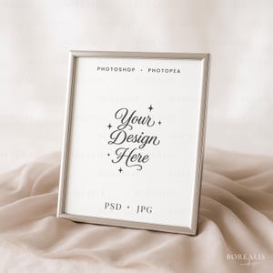 Könnte beinhalten: Ein silberner Bilderrahmen auf einer beigen Stoffoberfläche. Der Rahmen zeigt einen Druck mit dem Text "Your Design Here" in eleganter Schrift. Die Wörter "PHOTOSHOP + PHOTOPEA" und "PSD + JPG" sind ebenfalls auf dem Druck zu sehen.