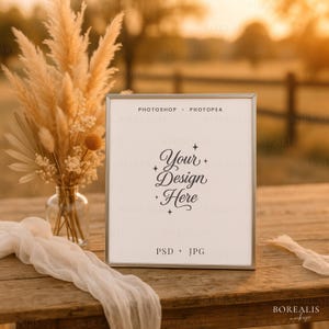 Könnte beinhalten: Ein quadratischer silberner Rahmen-Mockup mit dem Text "Your Design Here" in eleganter Schrift. Der Rahmen steht auf einem rustikalen Holztisch, mit einer Vase mit Trockenblumen und einem durchsichtigen weißen Stoff. Der Hintergrund ist verschwommen, mit warmem, goldenem Licht.