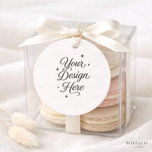May include: A clear, square gift box tied with a cream ribbon and bow, containing macarons in pink and beige. A round tag with the text "Your Design Here" is attached. The box is presented on a white surface.