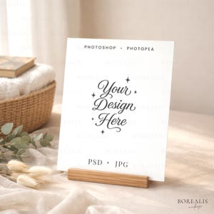 May include: A white card with the text "Your Design Here" in elegant script, held by a natural wood stand. The card also has the text "PHOTOSHOP + PHOTOPEA" and "PSD + JPG". The background includes a woven basket and soft fabric.