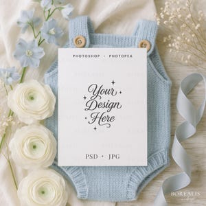 Peut inclure: Un body de bébé en tricot bleu clair avec des boutons en bois, tenant une carte blanche avec le texte "Your Design Here". La carte est entourée de fleurs blanches, de fleurs bleues et d'un ruban bleu clair. Le texte "PHOTOSHOP + PHOTOPAE" et "PSD + JPG" sont également visibles.