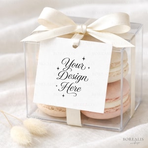 Könnte beinhalten: Eine transparente quadratische Box, gebunden mit einem cremefarbenen Band und einer Schleife, die zwei Makronen enthält. Ein weißes Etikett mit dem Text "Your Design Here" hängt am Band. Die Makronen sind hellrosa und cremefarben.