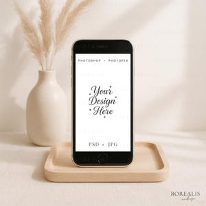 Könnte beinhalten: Ein schwarzes Smartphone mit transparenter Hülle liegt auf einem hellen Holztablett. Auf dem Bildschirm steht "Your Design Here" und "Photoshop + Photopea". Im Hintergrund eine weiße Vase mit getrocknetem Pampasgras.