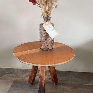 Op de afbeelding: Een ronde houten bijzettafel met drie donkere, getextureerde poten en een glad, lichtgekleurd rond blad. Op de tafel staat een doorzichtige, reliëf glazen fles gevuld met gedroogde bloemen en grassen, voorzien van een klein beige label.