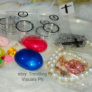 May include: A close-up of a variety of jewelry items, including a pearl bracelet with a pink charm, a silver chain necklace, and two sets of silver rings in different sizes. The rings are packaged in clear plastic bags with size labels. The jewelry is displayed on a white fuzzy surface.