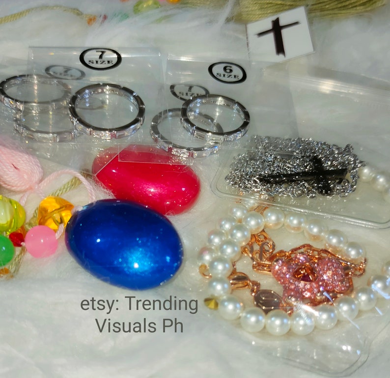 May include: A close-up of a variety of jewelry items, including a pearl bracelet with a pink charm, a silver chain necklace, and two sets of silver rings in different sizes. The rings are packaged in clear plastic bags with size labels. The jewelry is displayed on a white fuzzy surface.